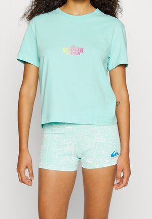Persona con camiseta azul claro de Stranger Things y pantalones cortos estampados a juego con el logo de Quiksilver en el muslo izquierdo.