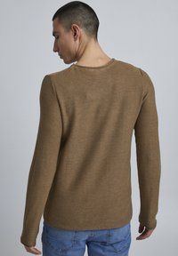 Maglione lungo marrone, testurizzato, con scollo a girocollo, maniche aderenti e una vestibilità rilassata. Indossato con jeans in denim blu.