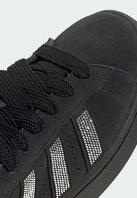 Crna antilop tenisica s ravnim crnim vezicama, ukrašena sjajnim srebrnim rhinestones duž tri pruge i teksturiranim gumenim potplatom.