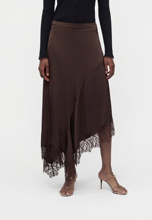 Femme portant une jupe asymétrique marron foncé avec bordure en dentelle, un haut noir à manches longues et des sandales à talons hauts à brides.