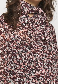 Kwiatowa bluzka z przezroczystej tkaniny na burgundowym tle, zdobiona małymi różowymi i białymi kwiatami, z kołnierzykiem na kokardkę i detalami z guzikami.