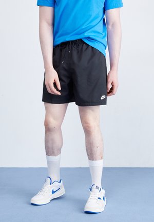 Personne portant un short Nike noir, un t-shirt bleu, des chaussettes blanches et des baskets blanches avec des touches bleues, debout sur un sol bleu clair.