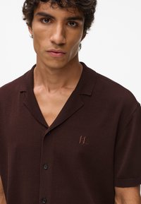 Tamsiai rudas trumpomis rankovėmis mygtukais užsegamas marškinėliai su minkšta tekstūra. Yra notch tipo apykaklė ir subtilus logotipas išsiuvinėtas ant krūtinės.