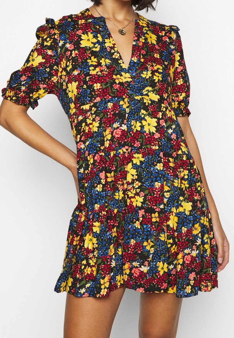 Femme portant une robe fleurie multicolore avec des fleurs jaunes, rouges et bleues, des manches courtes bouffantes et un ourlet à volants, posant avec une main sur la hanche.