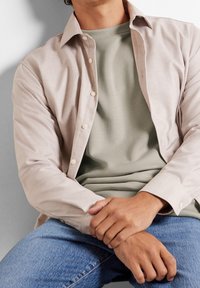 Camisa de botón de un beige claro con estampado, superpuesta a una camiseta verde apagado, combinada con jeans azules. La camisa tiene un corte relajado y botones visibles.
