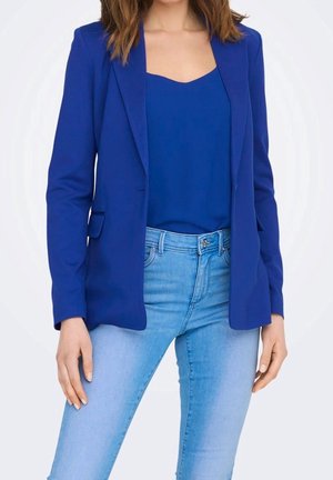 Femme portant un blazer bleu roi et un haut assorti avec un jean taille haute bleu clair, posant devant un fond blanc uni.