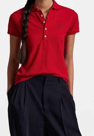 Woman with long braided hair wearing a red short-sleeve polo shirt and dark pleated trousers with hands in pockets.