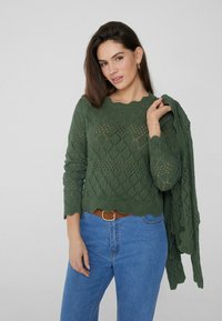 Grüner gestrickter Pullover mit Rautenmuster und gewelltem Saum, kombiniert mit einem passenden Schal. Verfügt über lange Ärmel und einen runden Halsausschnitt.