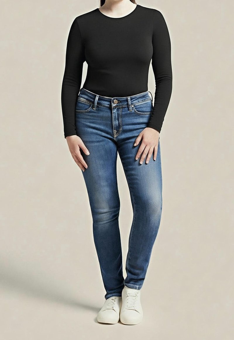 Personne portant un haut noir cintré à manches longues, un jean slim bleu avec des poches avant et des baskets blanches, debout devant un fond uni.