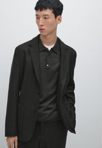 Dunkelblazer aus Wolle mit einer Knopfverschluss, zwei Vordertaschen, kombiniert mit einem dunklen Poloshirt. Glatte Textur und taillierte Passform.