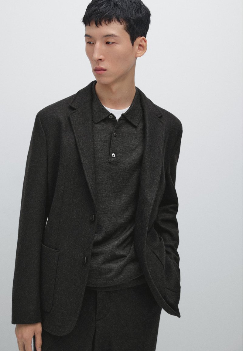 Dunkelblazer aus Wolle mit einer Knopfverschluss, zwei Vordertaschen, kombiniert mit einem dunklen Poloshirt. Glatte Textur und taillierte Passform.