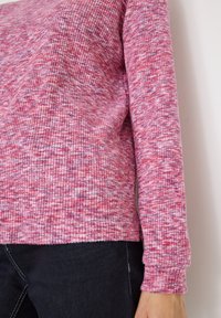 Różowy sweter robiony na drutach z fakturowanym, prążkowanym wzorem. Posiada długie rękawy i okrągły dekolt. Połączony z ciemnymi dżinsami.