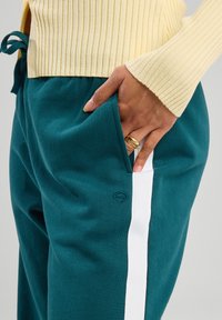 Pantaloni tuta turchesi con una striscia laterale bianca e logo ricamato, realizzati in un tessuto morbido. Una mano è appoggiata nella tasca laterale, rivelando un anello.