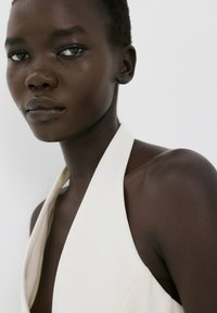 Primer plano de una persona de piel oscura y cabello corto, con un top blanco tipo halter, mirando ligeramente hacia otro lado sobre un fondo claro y liso.