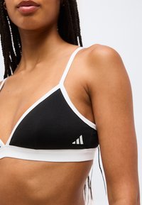 Γυναίκα που φοράει αθλητικό σουτιέν σε ασπρόμαυρο χρώμα με λεπτές τιράντες και μικρό λευκό λογότυπο στο αριστερό μέρος, μπροστά σε απλό φόντο.