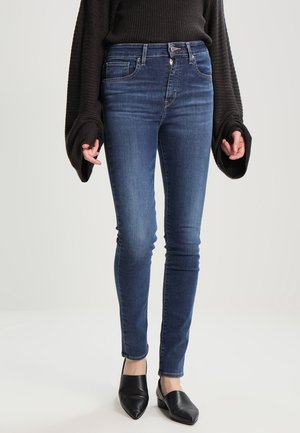 Vrouw draagt donkerblauwe skinny jeans, zwarte puntige schoenen en een zwarte geribde trui met uitgewaaierde mouwen.