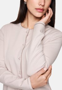 Hellrosa Strickjacke mit rundem Halsausschnitt, langen Ärmeln und Knopfverschluss. Funkelnde Akzente entlang der Knopfleiste und Ärmelenden. Weicher Textil.