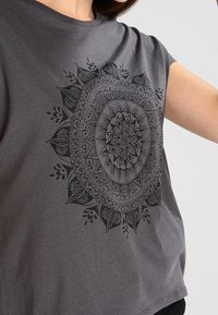 Šedé tričko s krátkým rukávem se černým mandala potiskem na přední straně, volný střih a hladká bavlněná textura.