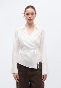 Λευκό πουκάμισο τύπου wrap με μακριά μανίκια, γιακά και δέσιμο στη μέση. Κατασκευασμένο από ελαφρύ ύφασμα με ομαλή υφή και άνετη εφαρμογή, συνδυάζεται με καφέ παντελόνια.