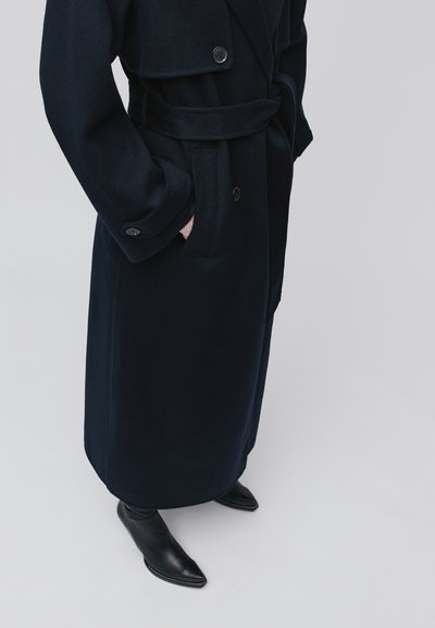 Granatowy płaszcz trench z wełny z dodatkiem, z paskiem w talii, dużymi klapami i dwoma guzikami. W zestawieniu z czarnymi skórzanymi botkami za kostkę o spiczastym nosku.