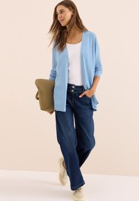 Vrouw die een lichtblauwe cardigan, witte top, donkerblauwe jeans en beige sneakers draagt, terwijl ze een olijfgroen kussen vasthoudt en tegen een effen achtergrond loopt.