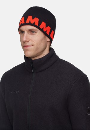 Bonnet en tricot noir avec le texte "MAMMUT" en orange vif. Texture douce, coupe ajustée. Porté avec une veste en polaire foncée dotée d'une fermeture éclair et d'un accent de logo.