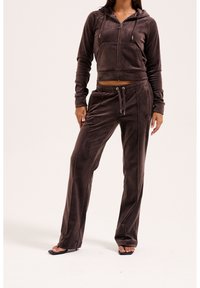Femme portant un sweat à capuche zippé en velours marron foncé et un pantalon assorti avec cordon de serrage, ainsi que des sandales noires à bouts ouverts sur un fond clair.