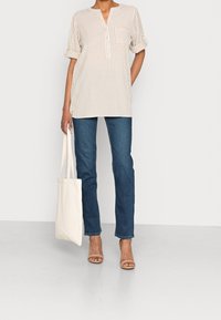 Camicia a righe beige chiaro con scollatura a V e taschino, abbinata a jeans in denim blu e una borsa a tracolla color crema. Modella che indossa sandali beige con tacco.