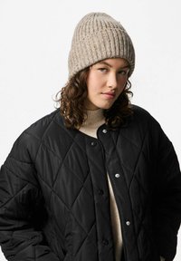 Giacca nera trapuntata con chiusure a pressione, berretto lavorato a maglia beige e dolcevita color crema. Presenta una texture liscia e un design caldo.