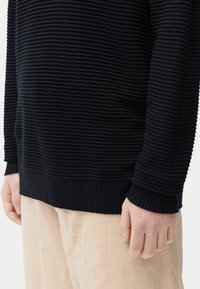 Pull noir côtelé avec des rayures horizontales texturées, comprenant un ourlet et des poignets côtelés épais. Associé à un pantalon beige clair.