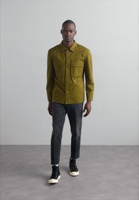 Olivengrøn skjorte med to brystlommer, parret med grå jeans og sorte sneakers, sat over en neutral baggrund.