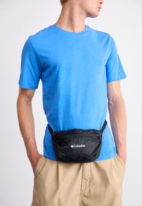 Pachet de talie negru Columbia cu închidere prin fermoar, curea ajustabilă și logo afișat proeminent, purtat peste un tricou albastru și pantaloni scurți bej.