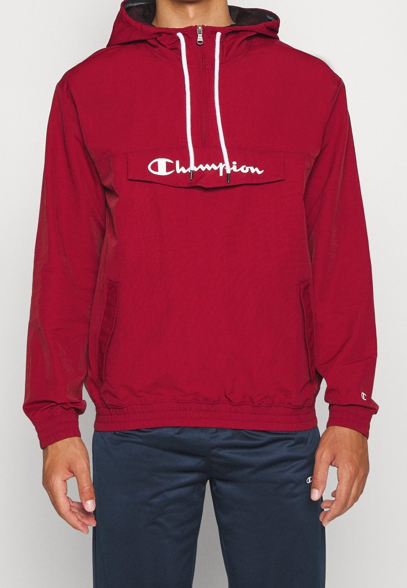 Coupe-vent rouge à capuche avec poignets et ourlet élastiques, doté d'une poche frontale et du logo blanc "Champion" sur la poitrine. Tissu léger.