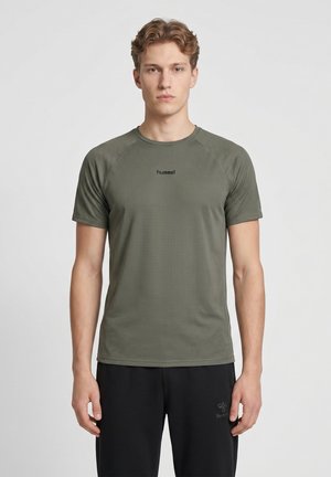 Jeune homme portant un t-shirt de sport vert olive à manches courtes et un pantalon de sport noir, debout devant un fond clair uni.
