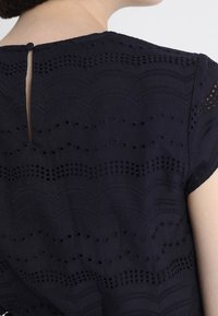 Nainen, jolla on päällään tummansininen pitsipusero, jossa on aaltomainen kuvio ja nappisuljin takana kaula-aukossa.