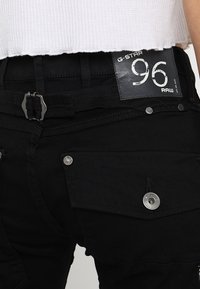 Černé kalhoty G-Star RAW s texturovanou tkaninou, kovovými detaily a štítkem s číslem 96. Obsahují kapsy a opasek.