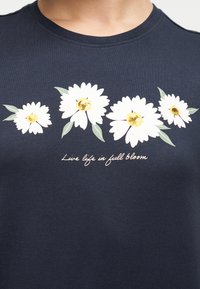 Μπλε ναυτικό πουκάμισο με τέσσερα λευκά μαργαρίτες με χρυσούς πυρήνες και πράσινα φύλλα, πάνω από το κείμενο «Ζήσε τη ζωή σε πλήρη άνθιση» σε καλλιγραφική γραφή.