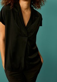 Schwarze Bluse mit kurzen Ärmeln, einem geknöpften Kragen und einem drapierten Frontdetail. Getragen mit schwarzen Hosen vor einem türkisfarbenen Hintergrund.