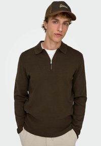 Brązowy sweter z dzianiny z kołnierzem na zamek, w połączeniu z beżową bluzą pod spodem. Brązowa czapka z napisem "Detroit" w złotych literach z przodu.