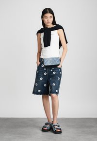 Hvid ærmeløs top, mørke denimshorts med blomsterbroderi, sort sweater draperet over skuldrene og blå mønstrede sandaler.