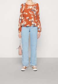 Rostfarbige Bluse mit langen Ärmeln und Peplum-Taille, kombiniert mit hellblauen Schlagjeans und braunen Sneakers. Kleine rosafarbene Handtasche.