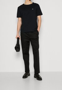 Maglietta nera a girocollo con un piccolo emblema, abbinata a pantaloni neri a gamba stretta. Uomo tiene una borsa nera a tracolla, indossando scarpe di pelle nere.