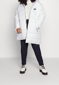 Biała puchowa kurtka z kapturem i zamkiem błyskawicznym, z czarnym logo Nike na piersi. W połączeniu z ciemnymi dżinsami i białymi butami na sznurowadła.