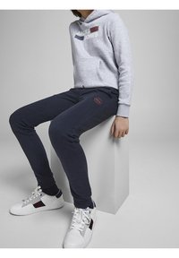 Šedá mikina s kapucí a přední kapsou, s logem "JACK & JONES", doplněná tmavě modrými zúženými tepláky s kulatou nášivkou. Bílé tenisky.