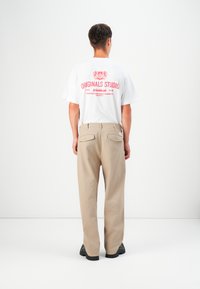 White cotton t-shirt with red graphic text on back, paired with beige pants, featuring back pockets and a straight-leg cut. Black footwear.