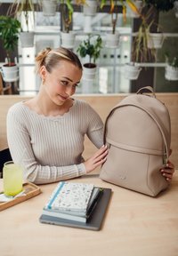 Beige backpack made of textured material, featuring a top handle, front zip pocket, and minimalistic design. Set on a wooden table.