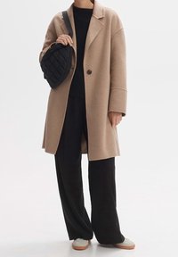 Manteau en laine beige avec une coupe décontractée et de grands revers, porté par-dessus un pull noir et un pantalon noir à jambes larges. Comprend un sac matelassé noir.