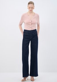 Cardigan corto de manga corta en rosa claro con botones, combinado con unos vaqueros de pierna ancha de color azul oscuro que cuentan con bolsillos frontales y una textura suave.