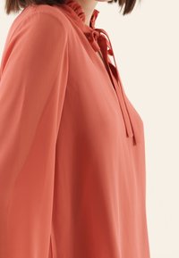 Blusa in corallo in tessuto leggero con colletto arricciato e dettaglio con laccio; presenta maniche lunghe e texture liscia.