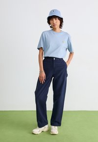 T-shirt en coton bleu clair avec un petit logo, pantalons larges bleus marine, chapeau seau en tricot bleu, et sneakers de sport crème.